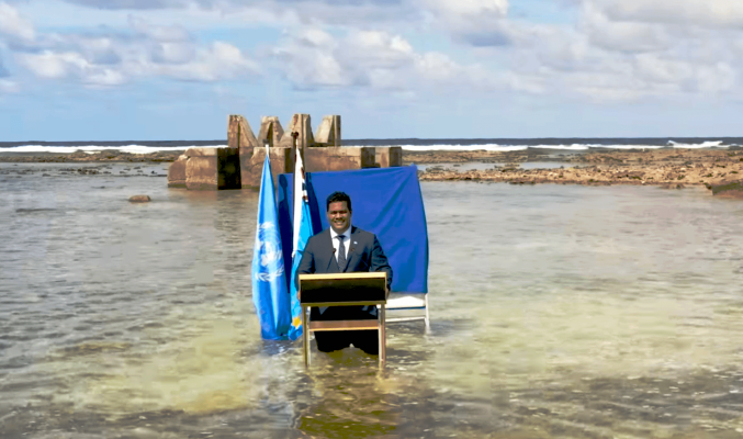 Ministr Tuvalu hovoří u řečnického pultíku o klimatu. Pultík stojí uprostřed moře.